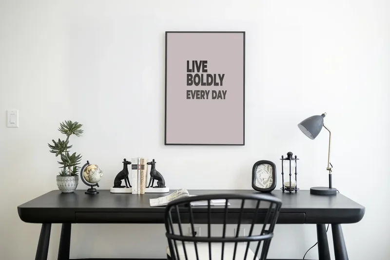 Framed "Live Boldly Every Day" typography print above a black desk in a minimalist home office with books, plants, a globe and a desk lamp.